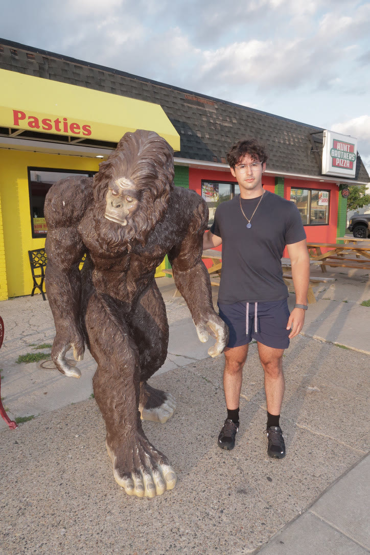Adam in a dark t-shirt standing beside a life-size Bigfoot statue outside a colorful roadside restaurant