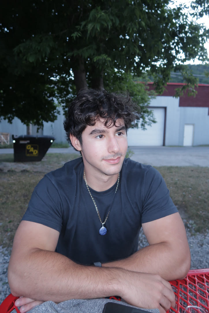 Close portrait of Adam at dusk in a dark t-shirt and stone pendant necklace, arms crossed, looking off to the side